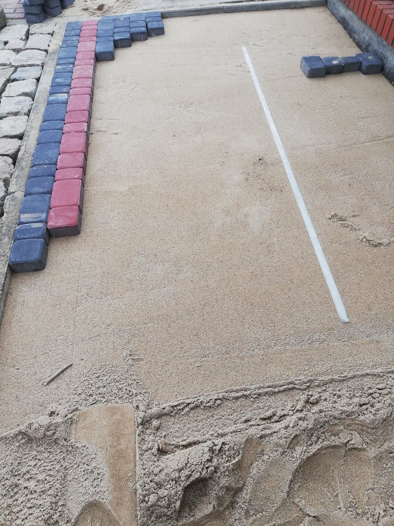 Układanie kostki brukowej: Widok z góry na przygotowany teren z podsypką piaskową, częściowo ułożoną kostką w kolorach czerwonym i grafitowym wzdłuż krawędzi oraz wyznaczoną linią prostą.