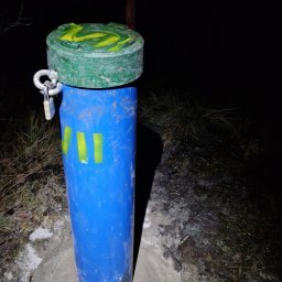 GEOLOG WIKTOR STRZELECKI - Niebieska rura osłonowa z zieloną pokrywą zabezpieczona kłódką, wystająca z betonowej podstawy na tle ciemnego terenu i nocy.