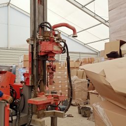 GEOLOG WIKTOR STRZELECKI - Wiertnica geologiczna wewnątrz hali magazynowej, wiercąca otwór w betonowej posadzce, obok stosy kartonów i wiader.