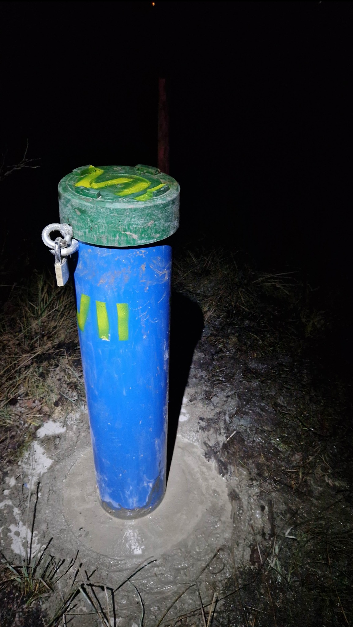 Niebieska rura osłonowa z zieloną pokrywą zabezpieczona kłódką, wystająca z betonowej podstawy na tle ciemnego terenu i nocy.