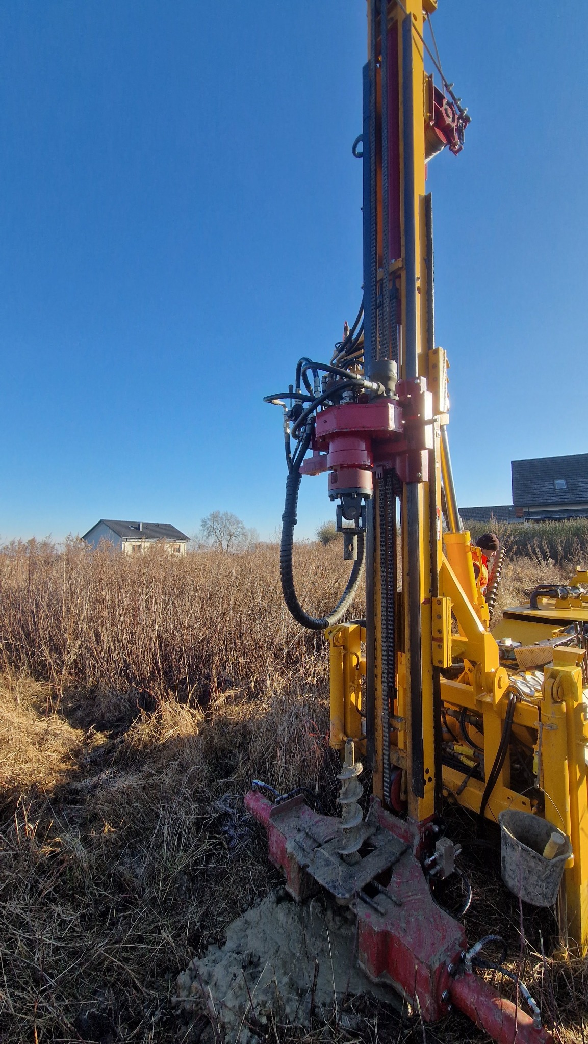 Żółto-czerwona wiertnica w trakcie pracy na polu, z widocznym świdrem w ziemi i budynkiem w tle pod błękitnym niebem.
