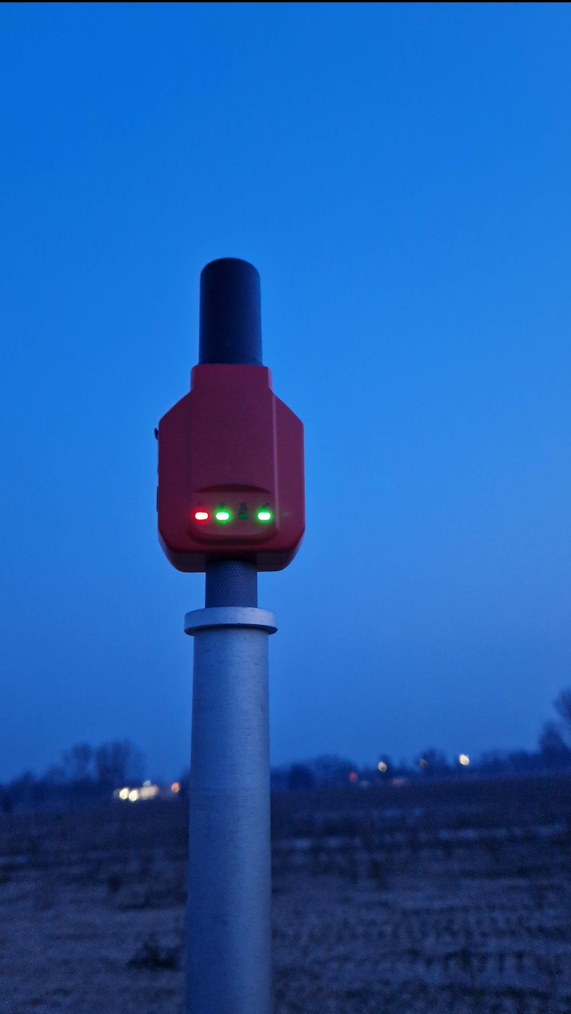 Czerwony odbiornik GNSS na metalowym maszcie, z trzema diodami LED (czerwona i dwie zielone) świecącymi na tle wieczornego nieba.