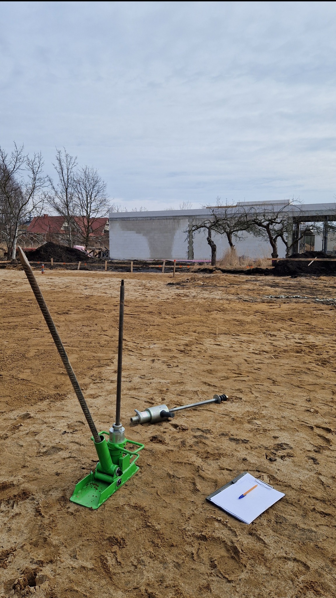 Badanie gruntu na placu budowy przy użyciu dynamicznej sondy lekkiej; widoczny sprzęt pomiarowy, notatnik i długopis.