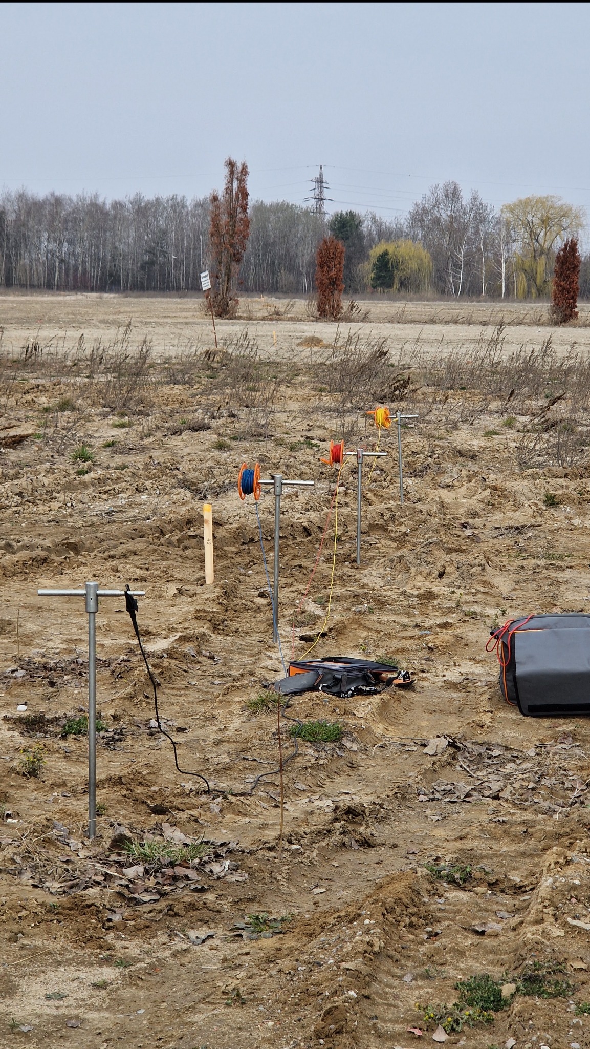 Badanie gruntu na polu: metalowe elektrody wbite w ziemię, połączone kolorowymi przewodami z aparaturą pomiarową w torbie. W tle widoczne drzewa i słup wysokiego napięcia.