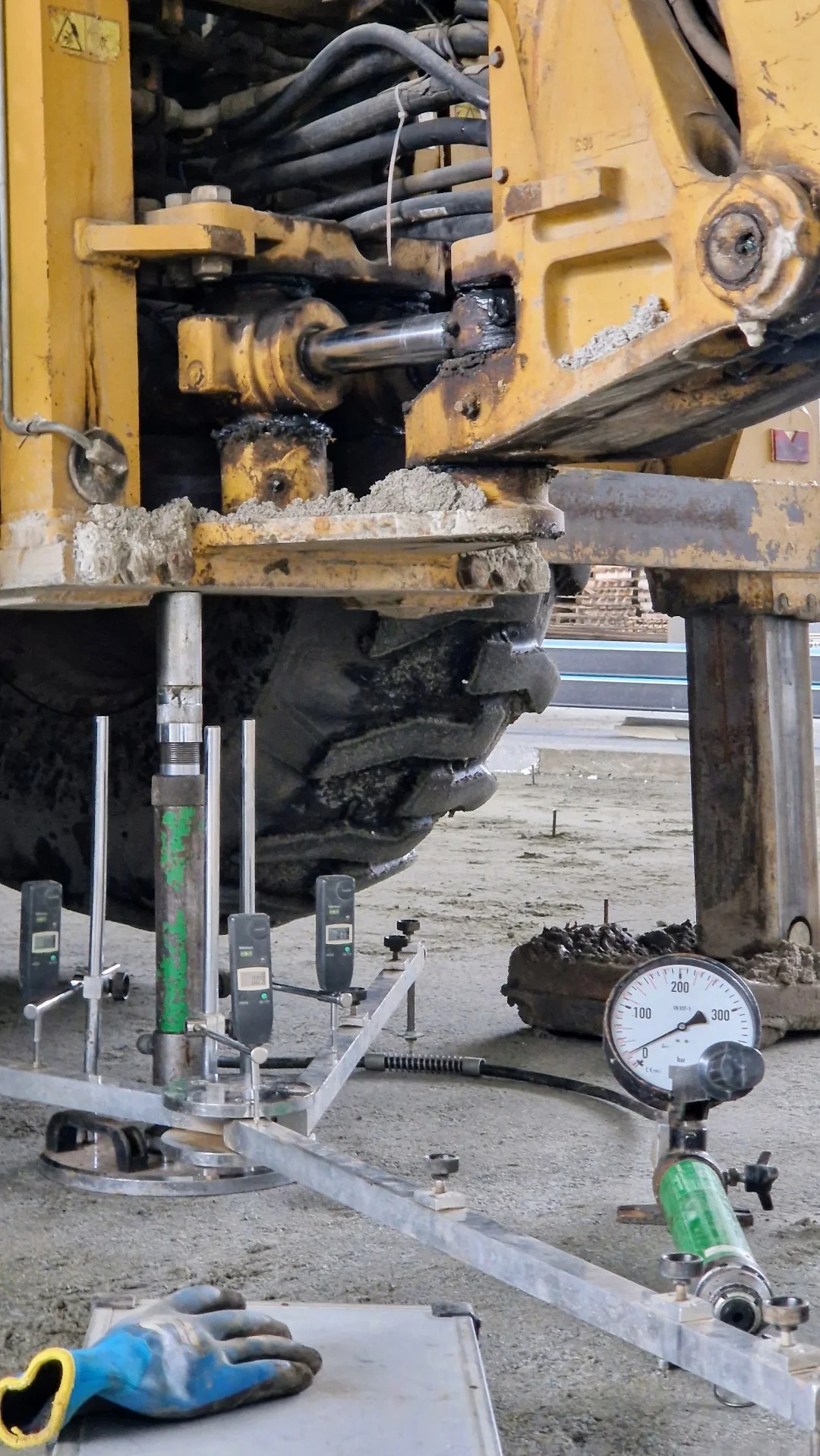 Badanie gruntu przy użyciu ciężkiego sprzętu z manometrem i czujnikami elektronicznymi, widoczne ubrudzone koło maszyny i niebieska rękawica robocza.