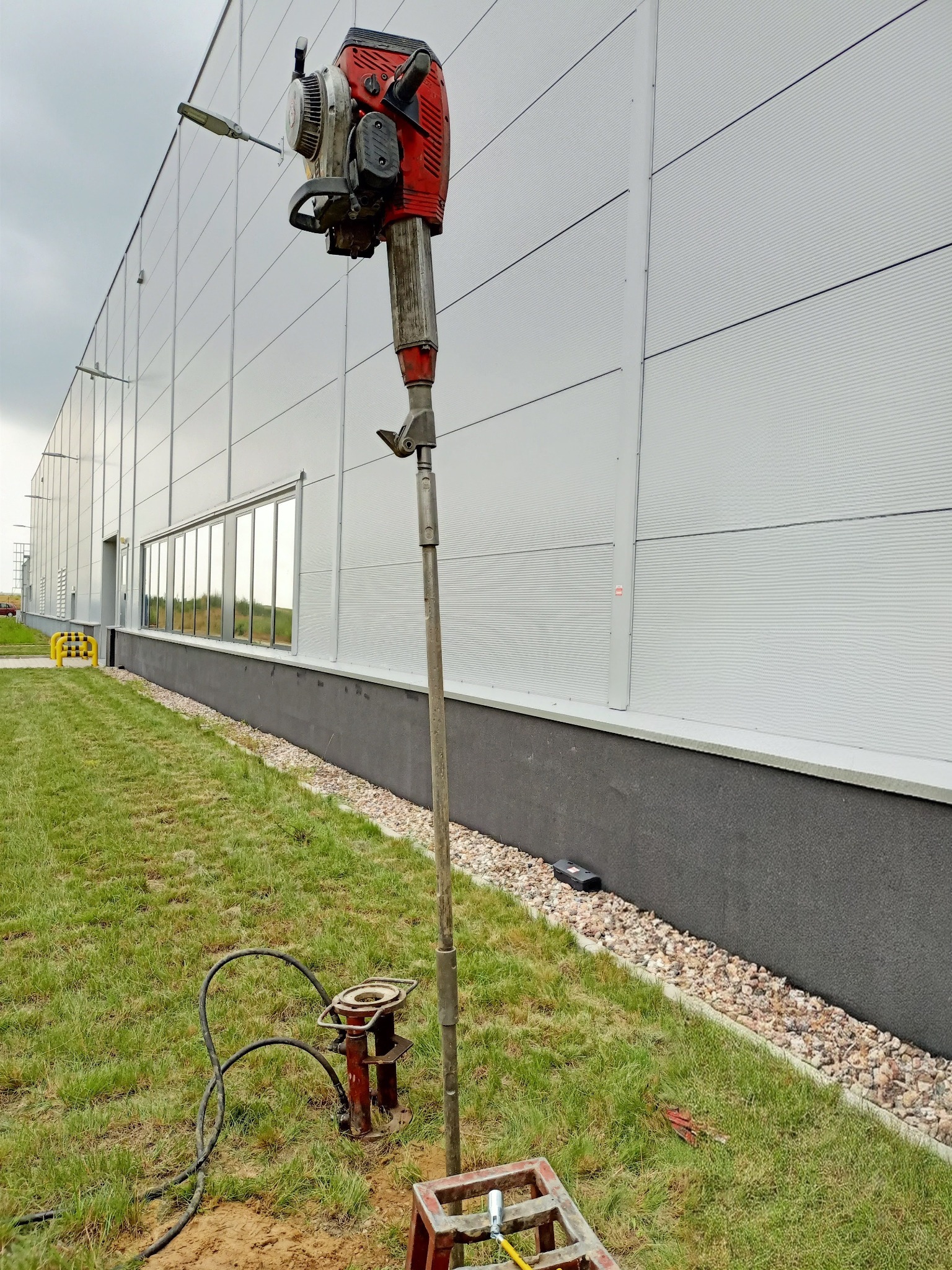 Czerwony spalinowy młot udarowy zamontowany na długim stalowym pręcie, w trakcie badania gruntu na trawiastym terenie przy budynku przemysłowym z szarą elewacją panelową.
