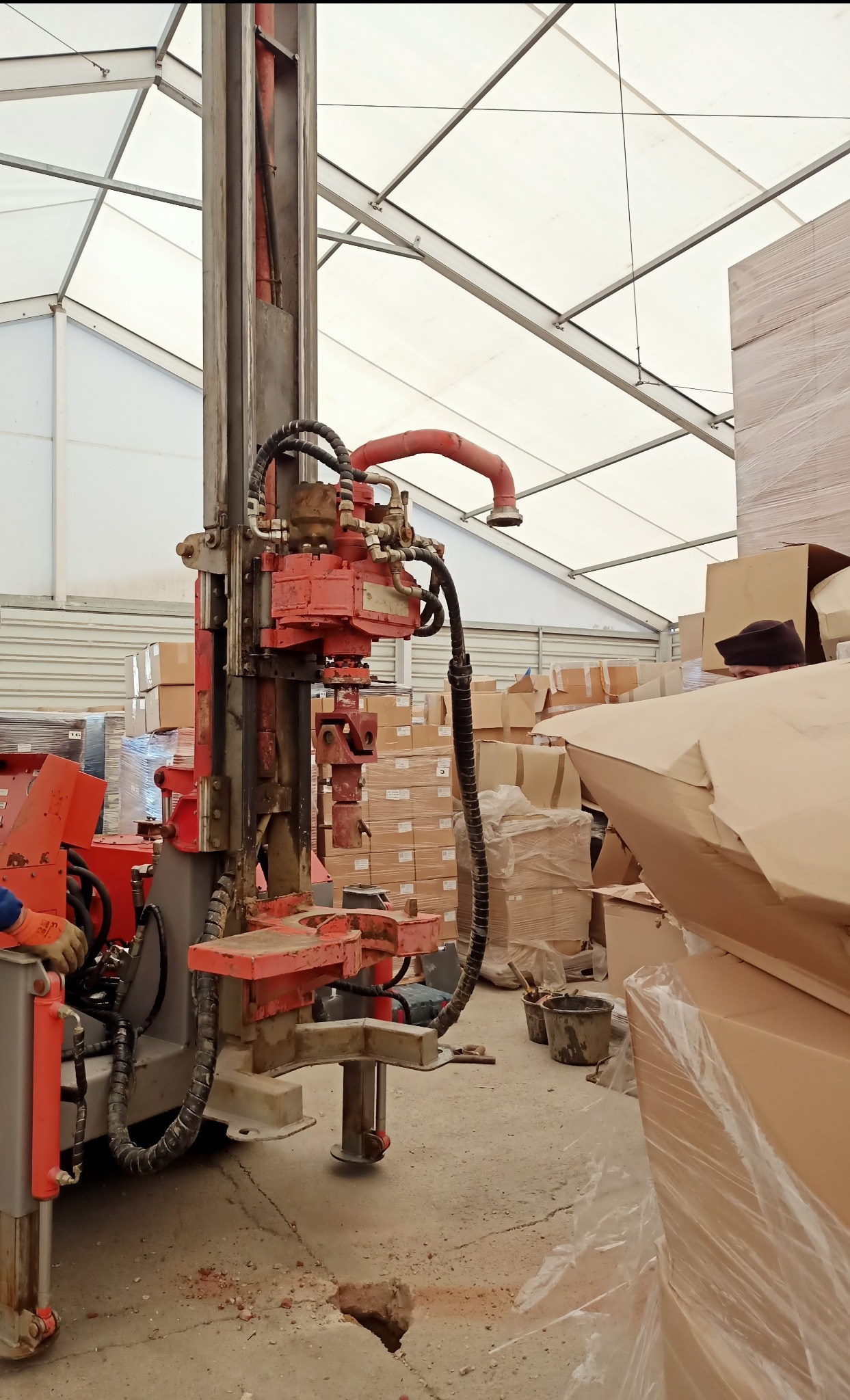Wiertnica geologiczna wewnątrz hali magazynowej, wiercąca otwór w betonowej posadzce, obok stosy kartonów i wiader.