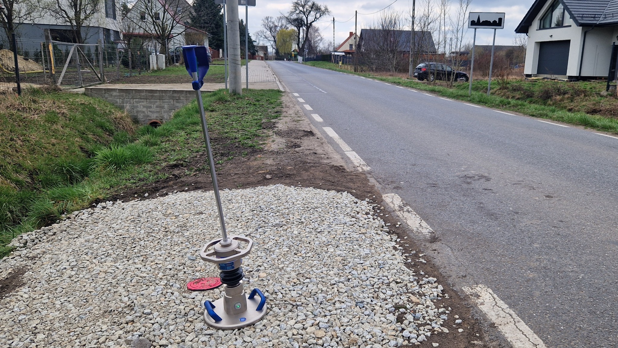 Urządzenie do zagęszczania gruntu stojące na żwirze przy drodze, w tle widoczna zabudowa wiejska i znak drogowy z sylwetką miasta.