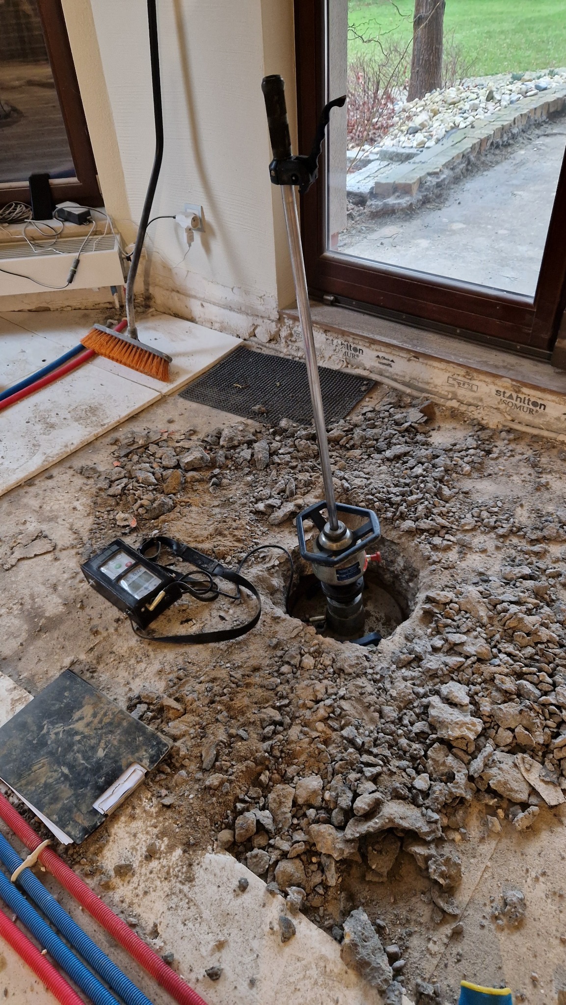 Wiercenie rdzeniowe w posadzce, widoczny wiertło rdzeniowe, gruz, przewody instalacyjne i przyrządy pomiarowe, okno w tle.