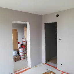 Wnętrze w trakcie prac wykończeniowych: świeżo pomalowane na szaro ściany z widocznymi otworami na instalacje, dwa surowe otwory drzwiowe, w tle widoczne puszki z farbą i kartony z materiałami...