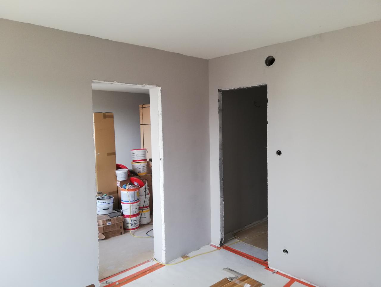 Wnętrze w trakcie prac wykończeniowych: świeżo pomalowane na szaro ściany z widocznymi otworami na instalacje, dwa surowe otwory drzwiowe, w tle widoczne puszki z farbą i kartony z materiałami...