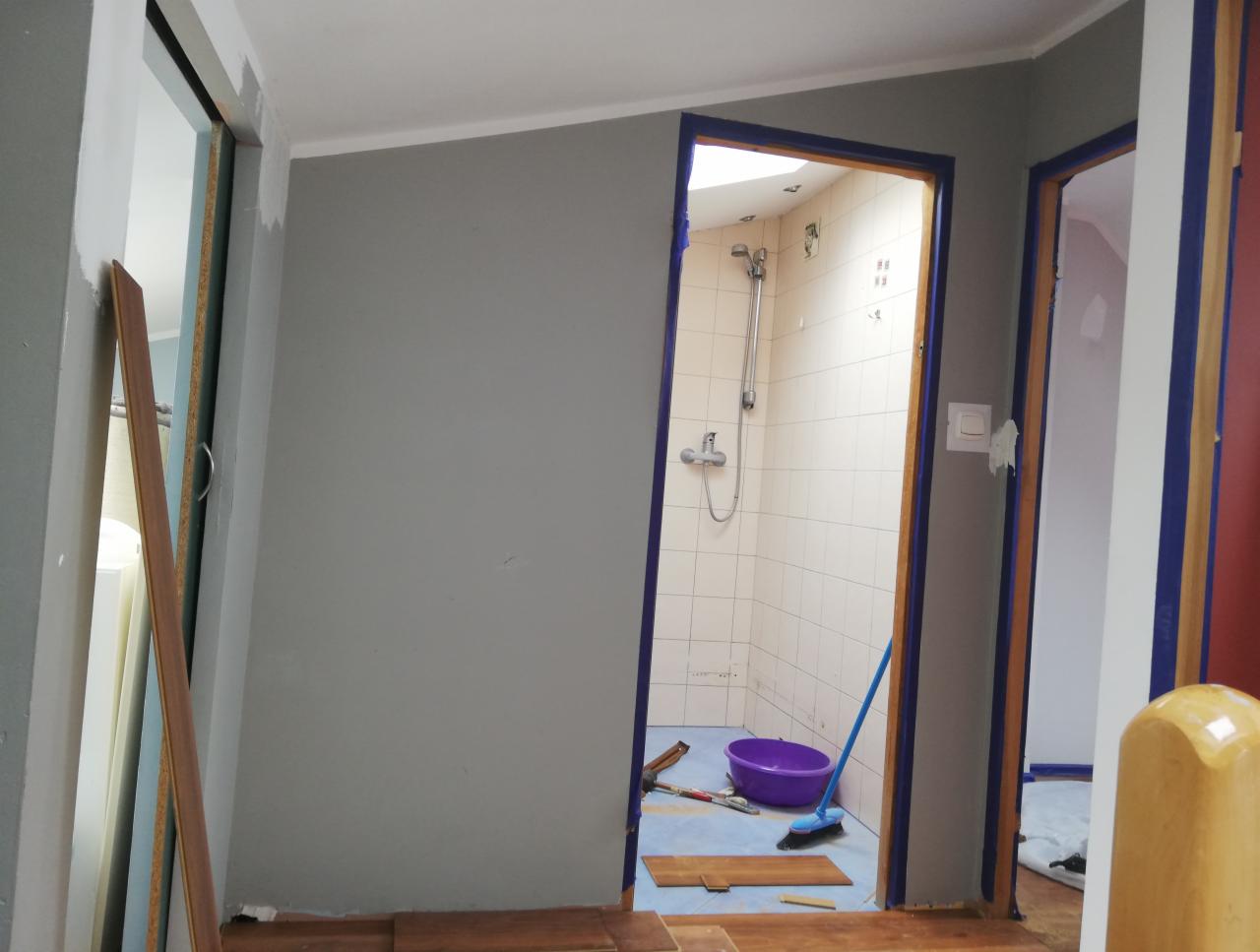 Wnętrze w trakcie remontu: szare ściany, fragment łazienki z prysznicem widoczny przez otwarte drzwi, narzędzia i wiadro na podłodze, listwy przypodłogowe oparte o ścianę.