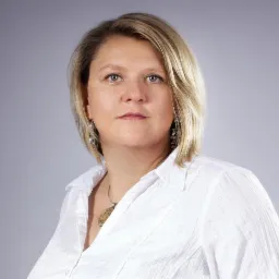 Portret kobiety z blond włosami w białej koszuli na szarym tle, z delikatnym makijażem i srebrną biżuterią.