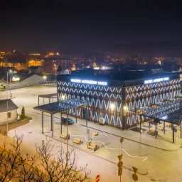 Nocne ujęcie z drona na oświetlony budynek dworca autobusowego w Sanoku z charakterystyczną elewacją w kształcie krzyży i zadaszeniami pokrytymi panelami słonecznymi, w tle miasto rozświetlone...