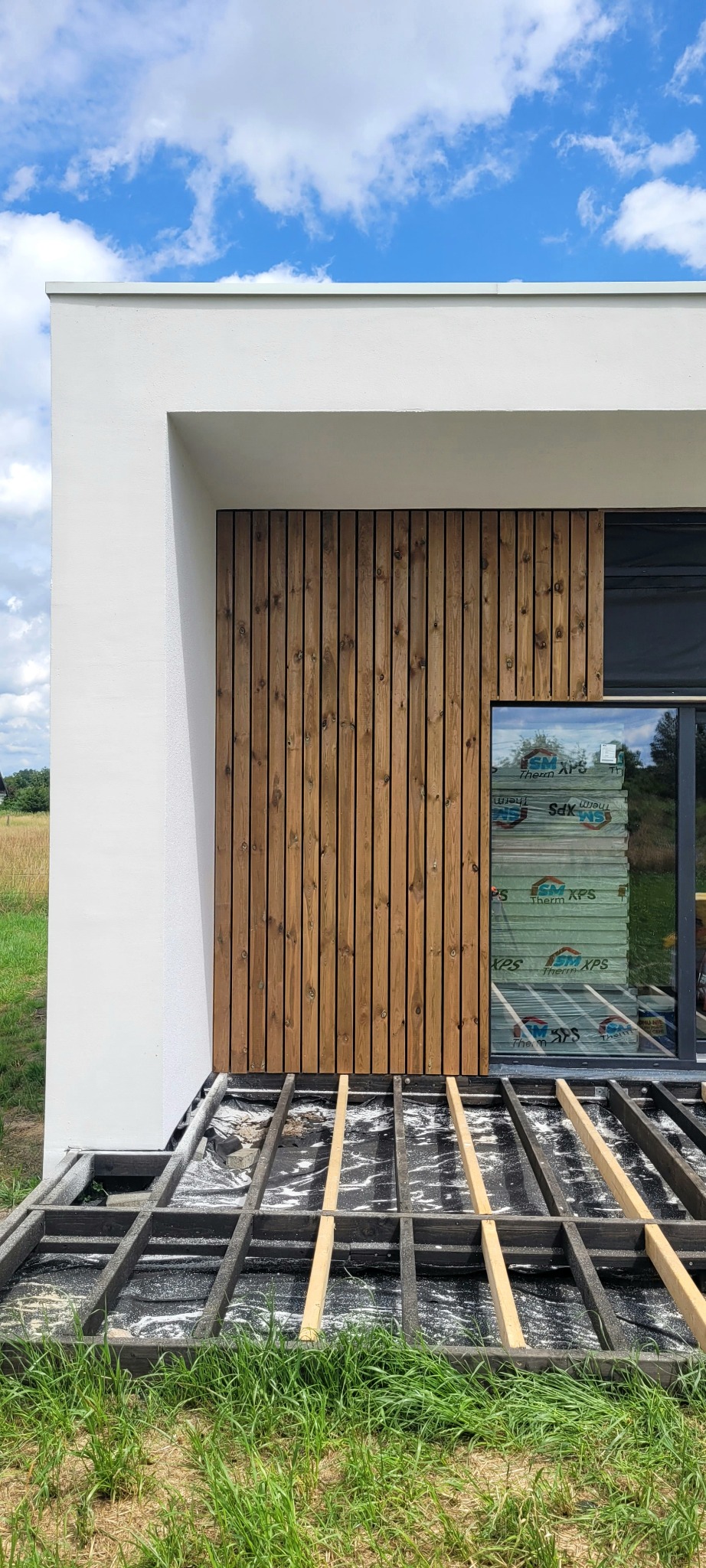 Nowoczesny dom szkieletowy w trakcie budowy: drewniana elewacja, widoczna konstrukcja podłogi, białe ściany. W oknie widoczne paczki izolacji.