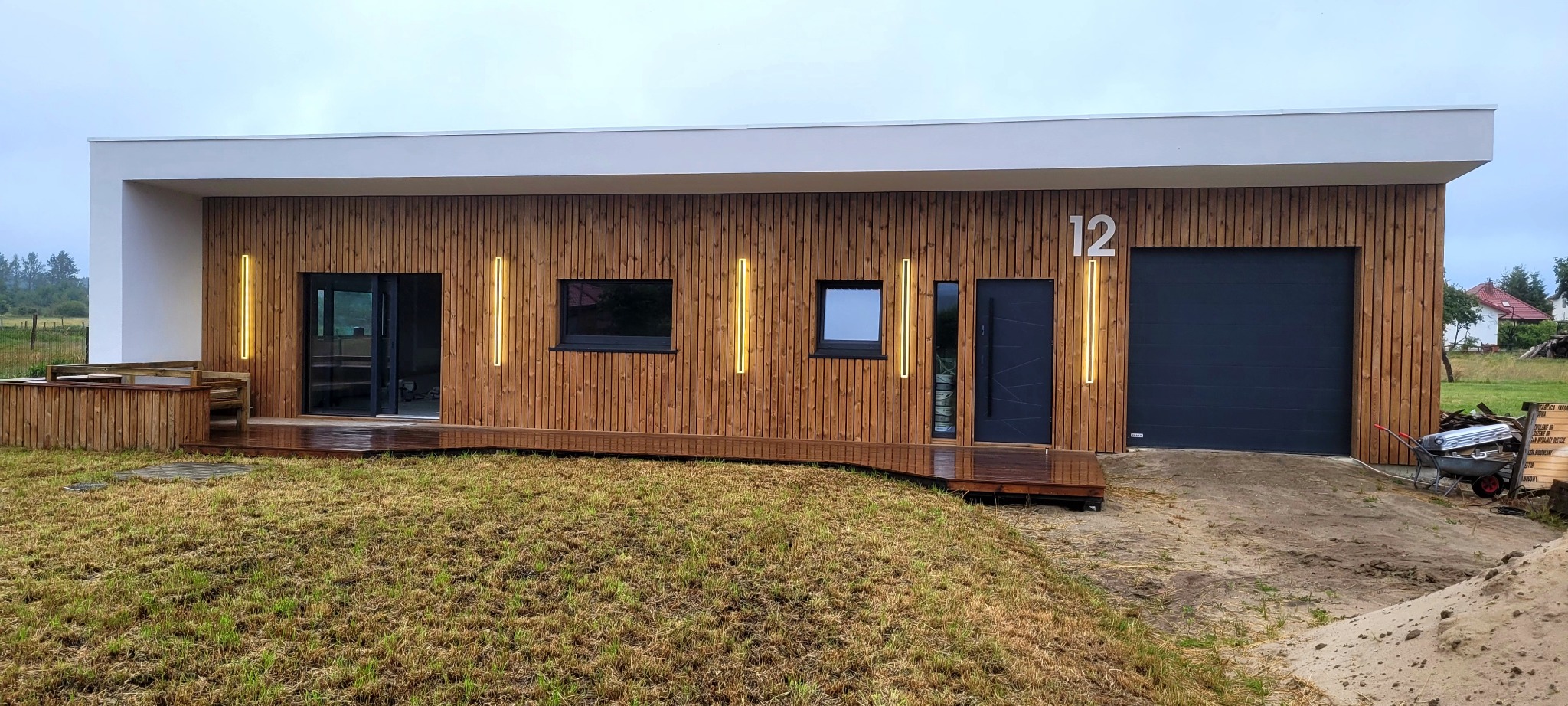 Nowoczesny dom szkieletowy z drewnianą elewacją, numerem 12 i oświetleniem LED. Widoczny taras i brama garażowa. Trawa w tle.
