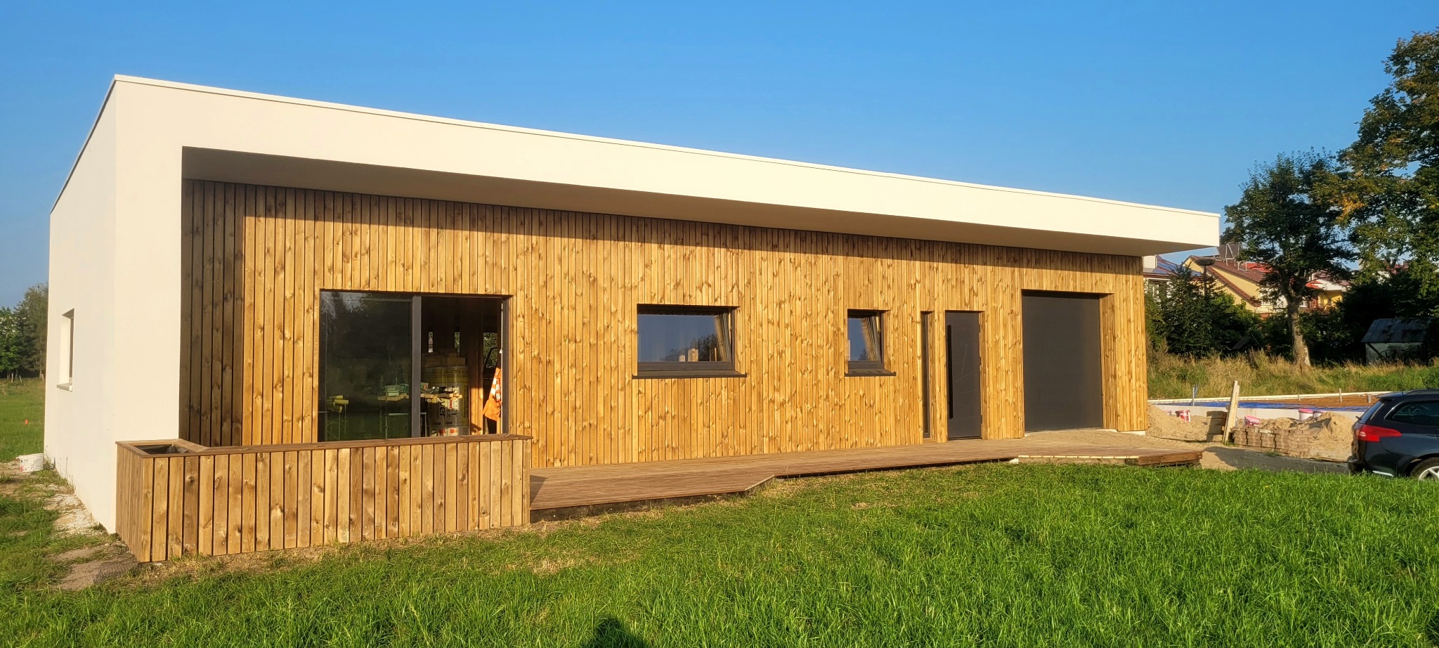 Nowoczesny dom szkieletowy z drewnianą elewacją i płaskim dachem, otoczony zielonym trawnikiem. Widoczne okna, drzwi wejściowe i fragment tarasu.