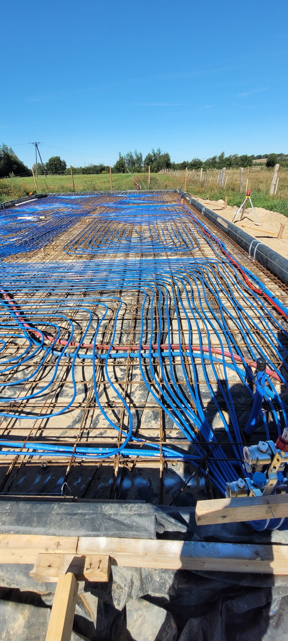 Instalacja ogrzewania podłogowego w domu szkieletowym: niebieskie rury ułożone na metalowej siatce z widocznym laserowym niwelatorem w tle, na tle zieleni.