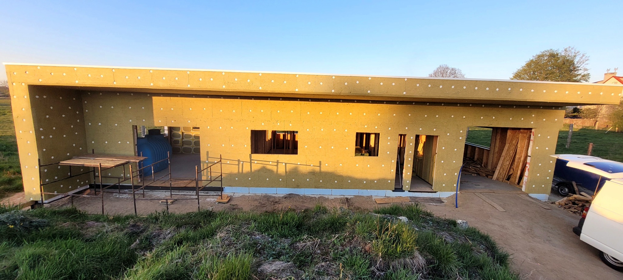 Budowa domu szkieletowego w trakcie: ocieplone ściany zewnętrzne z widocznymi otworami, rusztowanie, materiały budowlane. Ujęcie z zewnątrz, wczesny etap prac.