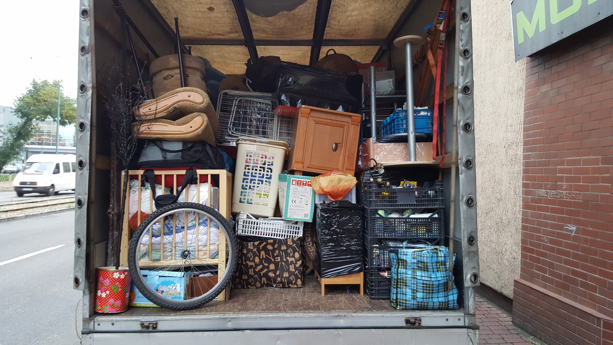 Wnętrze wypełnionej po brzegi furgonetki z meblami, skrzyniami i torbami, widoczne koło rowerowe, fragment ulicy z tramwajem w tle.