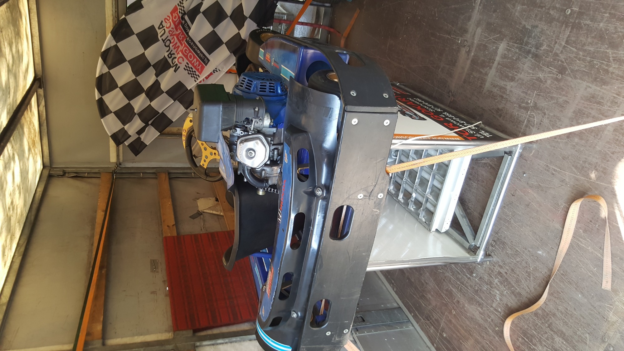 Go-kart zabezpieczony pasami w przestrzeni ładunkowej busa, widoczna flaga w szachownicę.