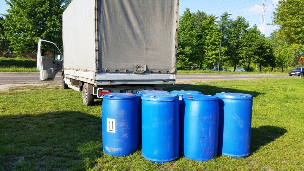 Biały samochód dostawczy z otwartymi drzwiami i sześć niebieskich beczek z tworzywa sztucznego stojących na trawie, przygotowanych do transportu.