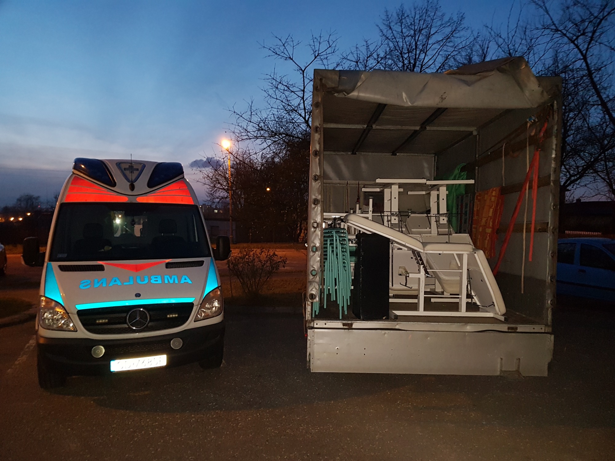 Ciężarówka z otwartą przestrzenią ładunkową, wypełniona meblami ogrodowymi, oraz zaparkowany obok ambulans Mercedes-Benz Sprinter w szaro-niebieskiej kolorystyce, sfotografowane o zmierzchu.