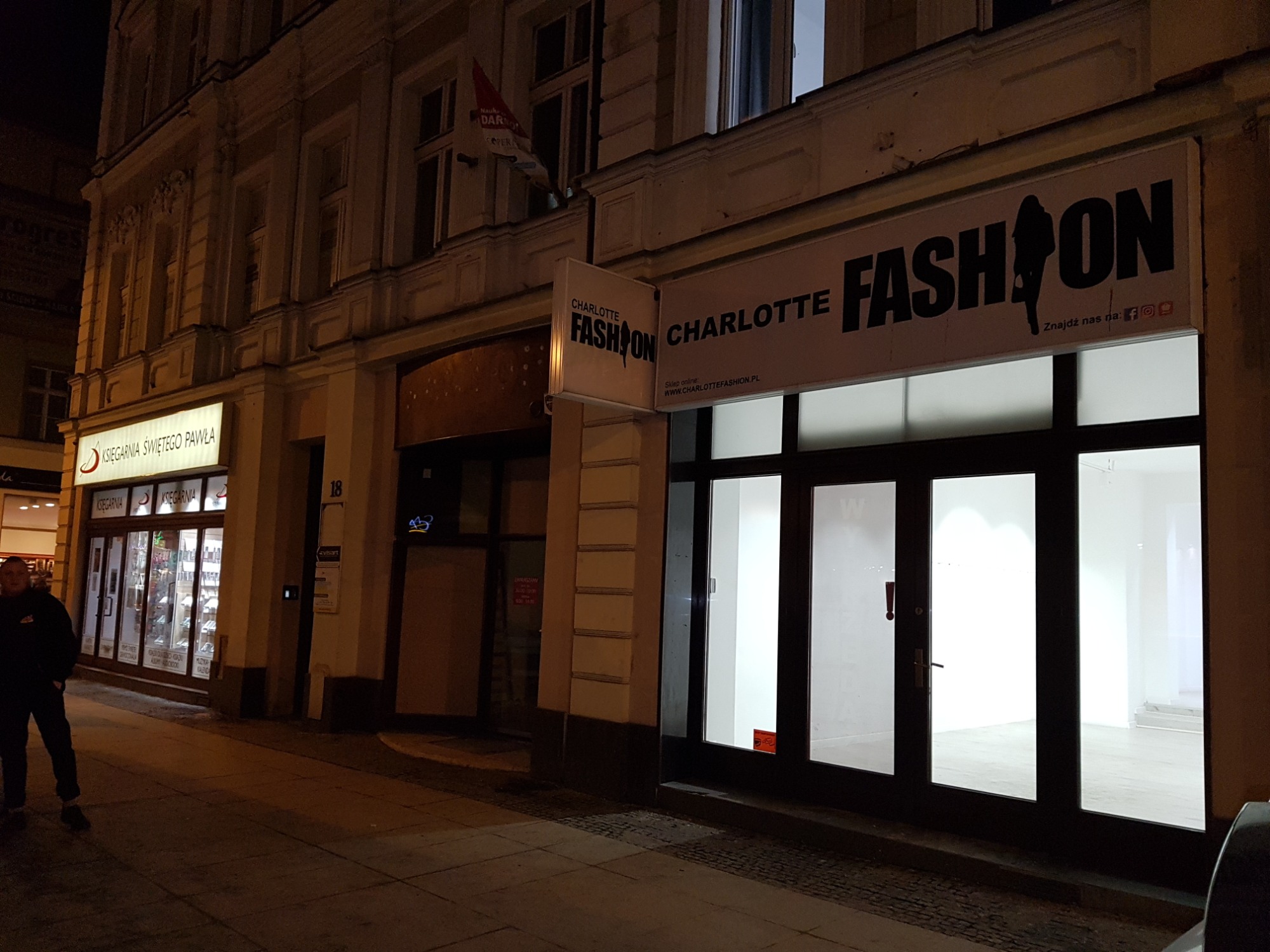 Nocny widok na fasadę kamienicy z szyldem 'CHARLOTTE FASHION' nad pustym lokalem, obok księgarnia i przechodzień w ciemnym ubraniu.