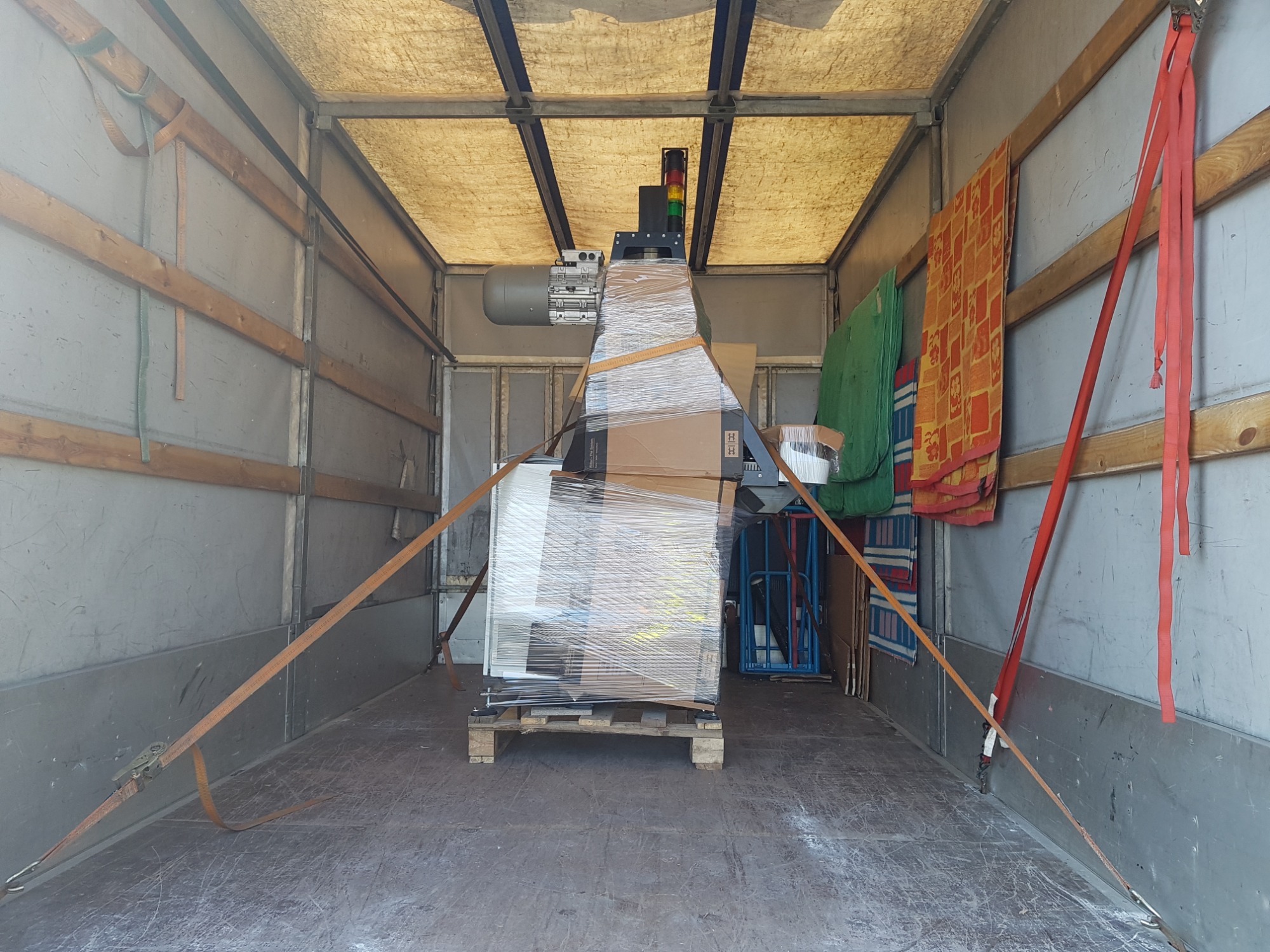 Maszyna przemysłowa owinięta folią stretch, zabezpieczona pasami transportowymi na palecie wewnątrz przestrzeni ładunkowej busa.