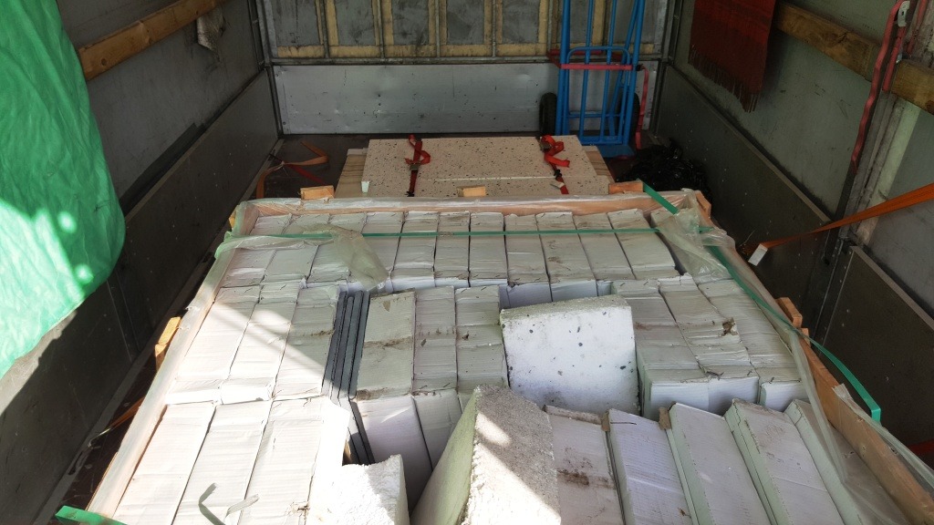 Wnętrze skrzyni ładunkowej wypełnionej paletami z materiałami budowlanymi, zabezpieczonymi pasami transportowymi, z widocznym wózkiem transportowym w tle.