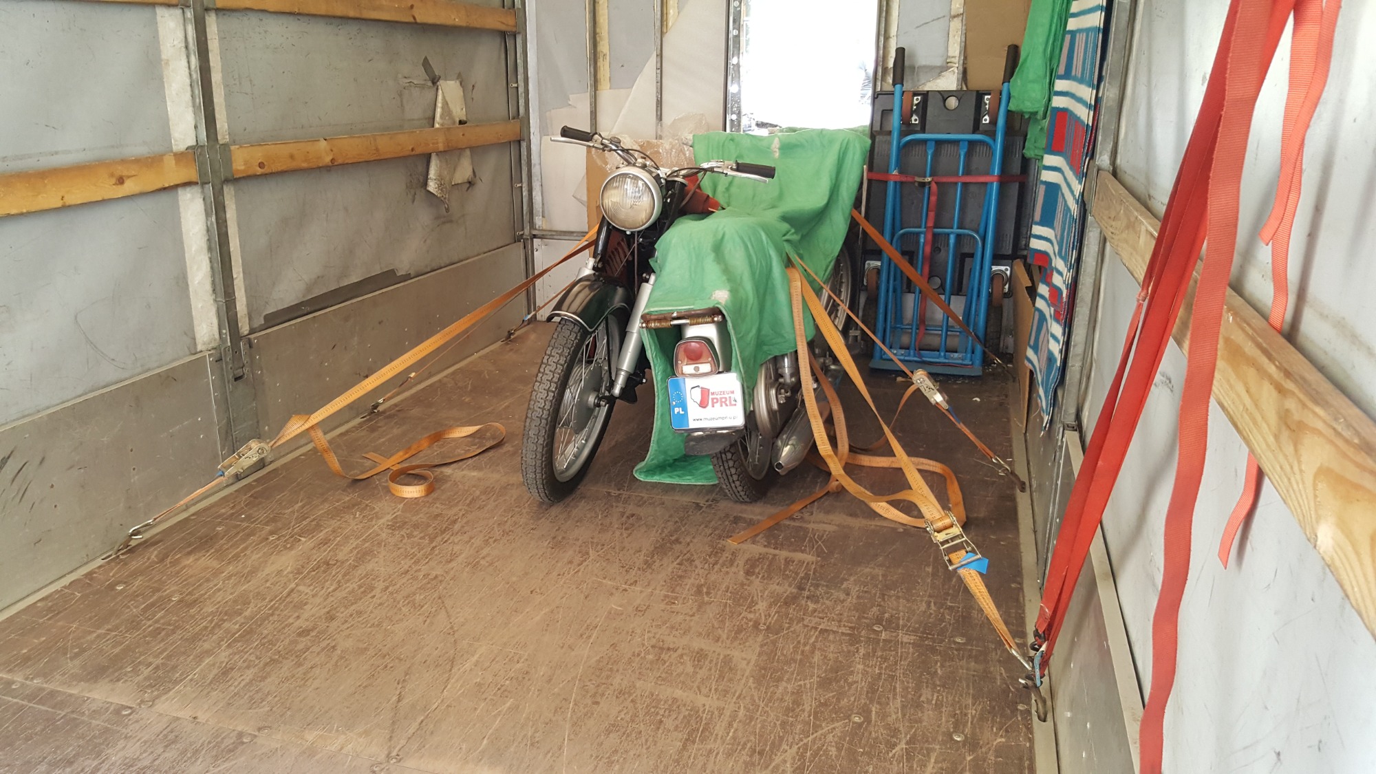 Motocykl Jawa TS 350 zabezpieczony pasami transportowymi wewnątrz przestrzeni ładunkowej busa, widoczny wózek transportowy i koc.