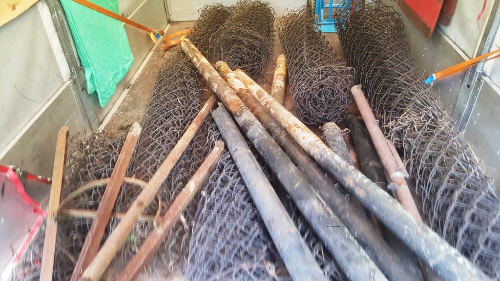 Wnętrze przestrzeni ładunkowej z metalowymi rolkami siatki ogrodzeniowej, rdzewiejącymi metalowymi słupkami i elementami ogrodzenia zabezpieczonymi pasami transportowymi.