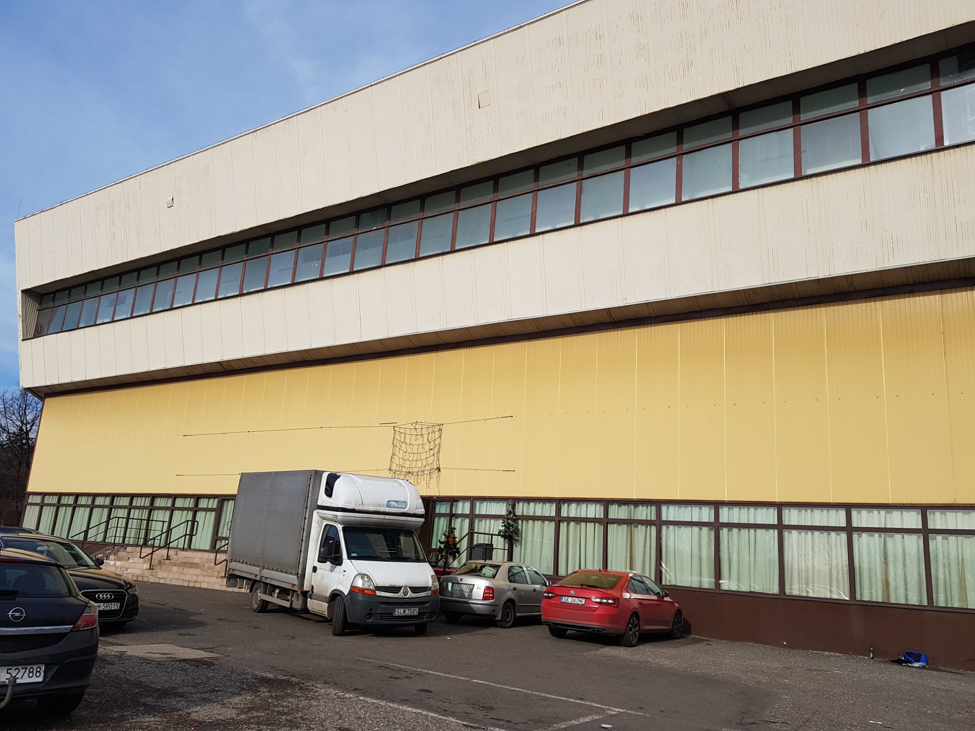 Biały samochód dostawczy zaparkowany przed dużym, żółtym budynkiem z rzędami okien, obok zaparkowane osobowe auta.