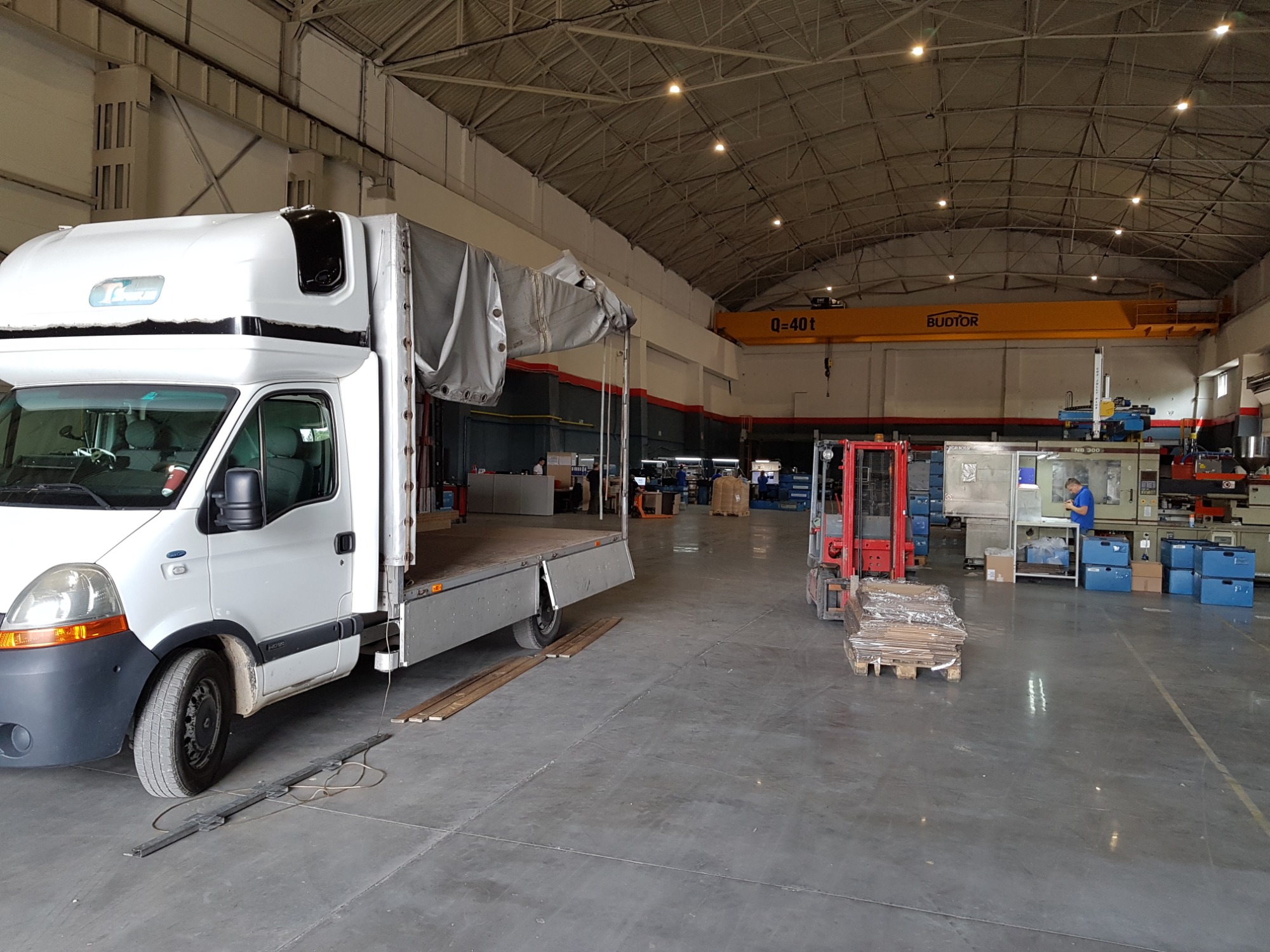 Biały samochód dostawczy z otwartą przestrzenią ładunkową w hali przemysłowej z suwnicą BUDTOR, w tle widać wózki widłowe i pracownika