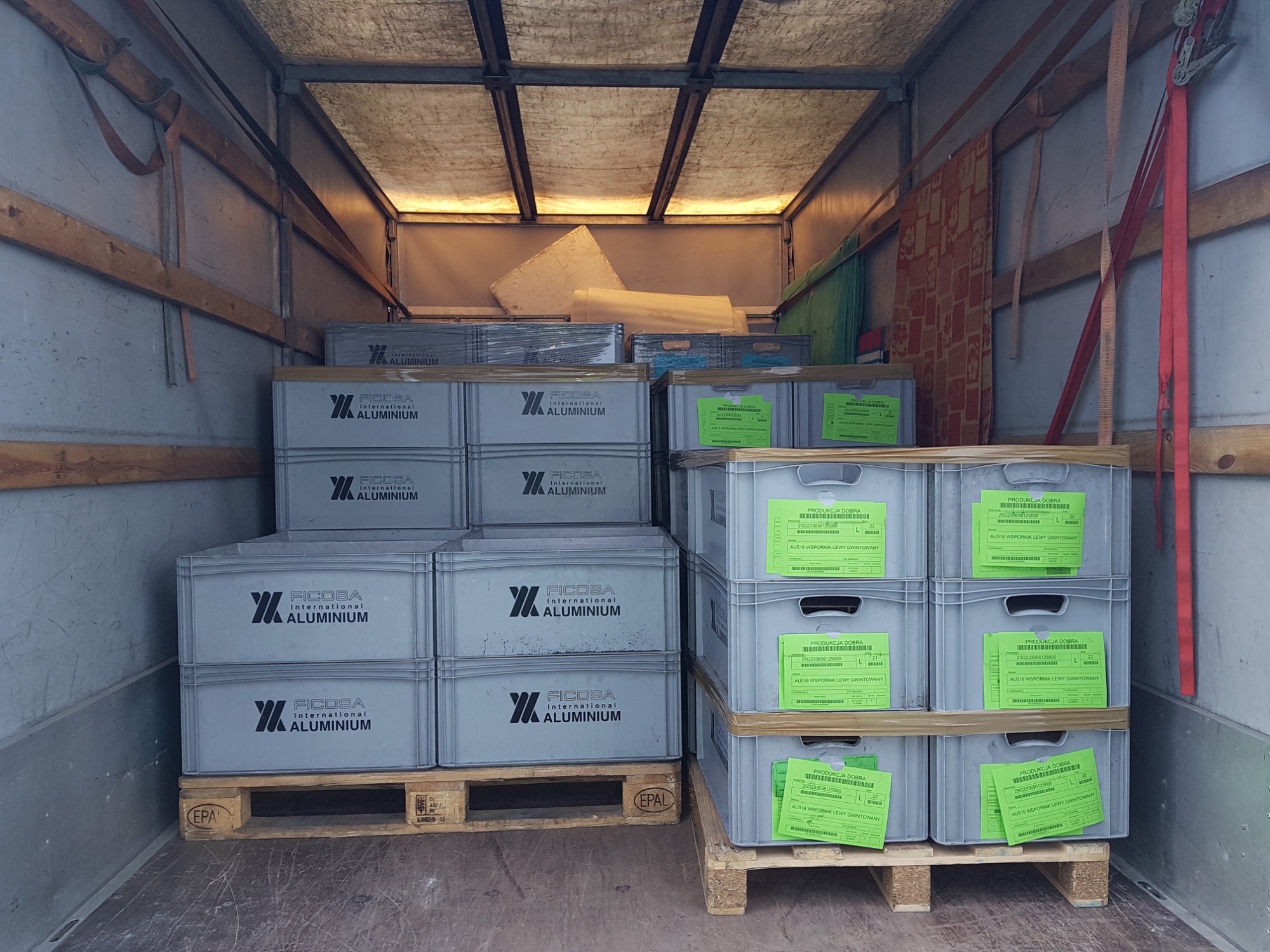 Wnętrze furgonetki załadowane szarymi plastikowymi skrzyniami z logo Ficosa International Aluminium, ułożonymi na drewnianych paletach, zabezpieczone pasami transportowymi.