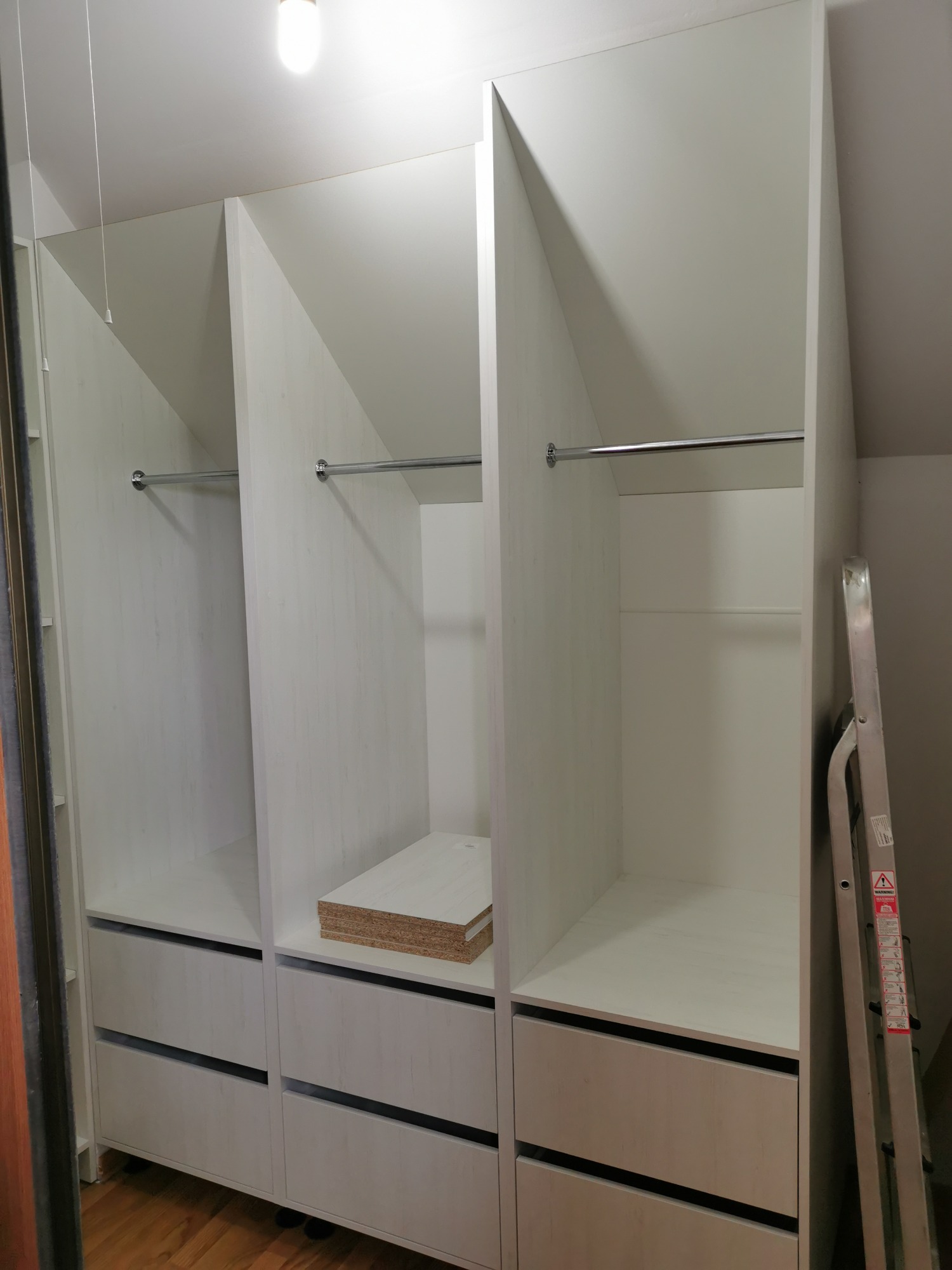 Wnętrze białej, częściowo zmontowanej szafy wnękowej z szufladami i drążkami na ubrania, widoczna drabina oparta o bok szafy.