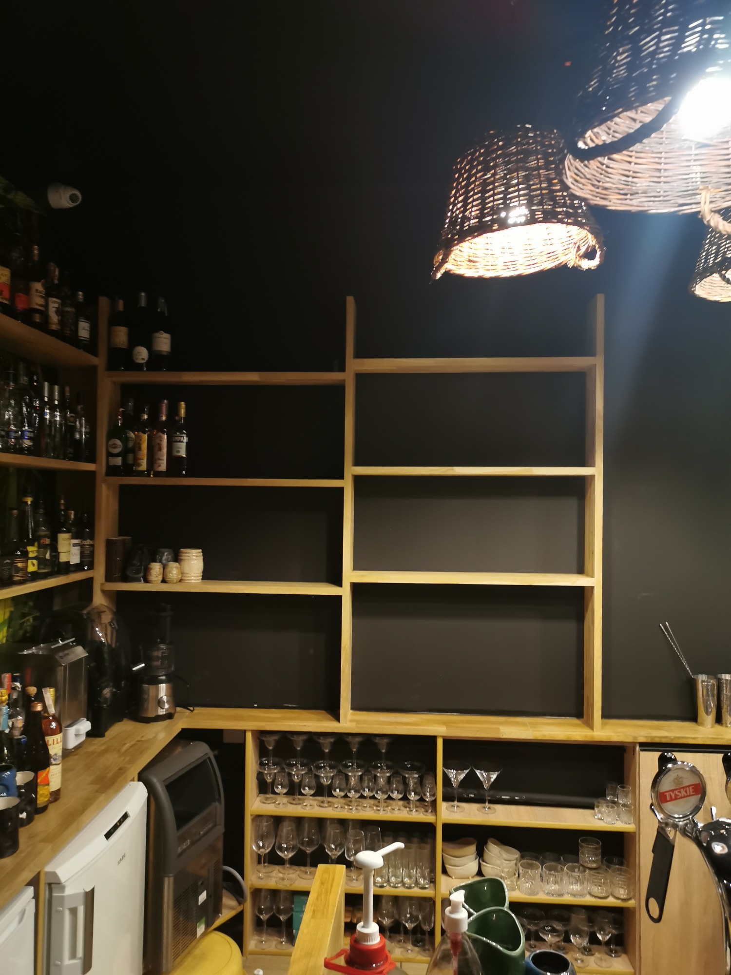 Półki barowe z jasnego drewna na tle ciemnej ściany, podświetlone wiklinowymi lampami, z widocznymi szklankami, butelkami i sprzętem barmańskim.