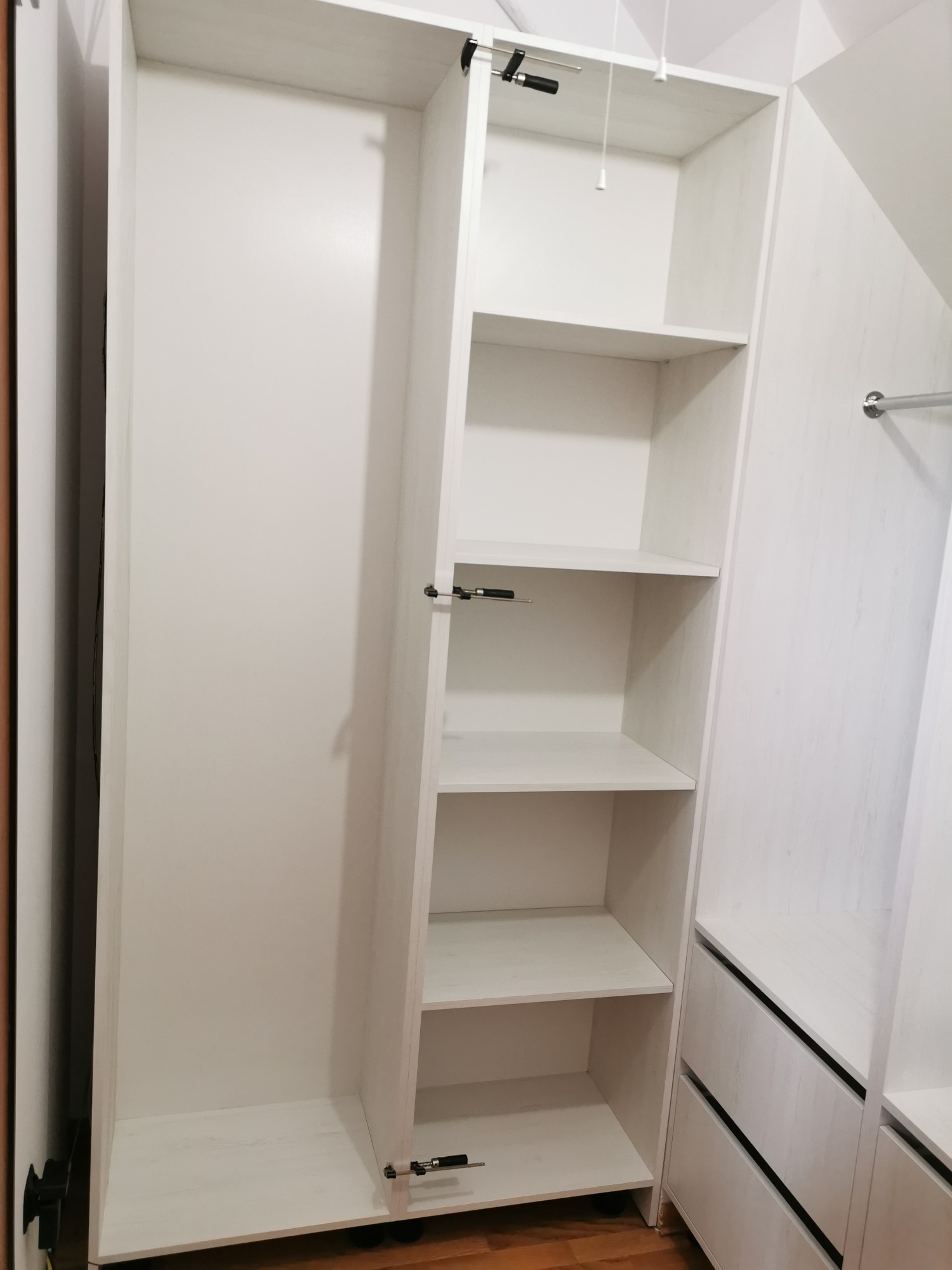 Wnętrze białej, drewnopodobnej szafy z półkami i szufladami, widoczne mechanizmy mocujące półki.