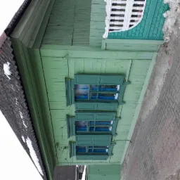 Zielony, drewniany dom z trzema oknami z ozdobnymi okiennicami, widoczne fragmenty dachu pokrytego eternitem i chodnika z kostki.