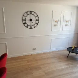 Salon po remoncie z drewnianą podłogą, białą ścianą z dekoracyjnymi listwami, zegarem, kinkietami, stolikiem z dekoracjami oraz zasłonami.