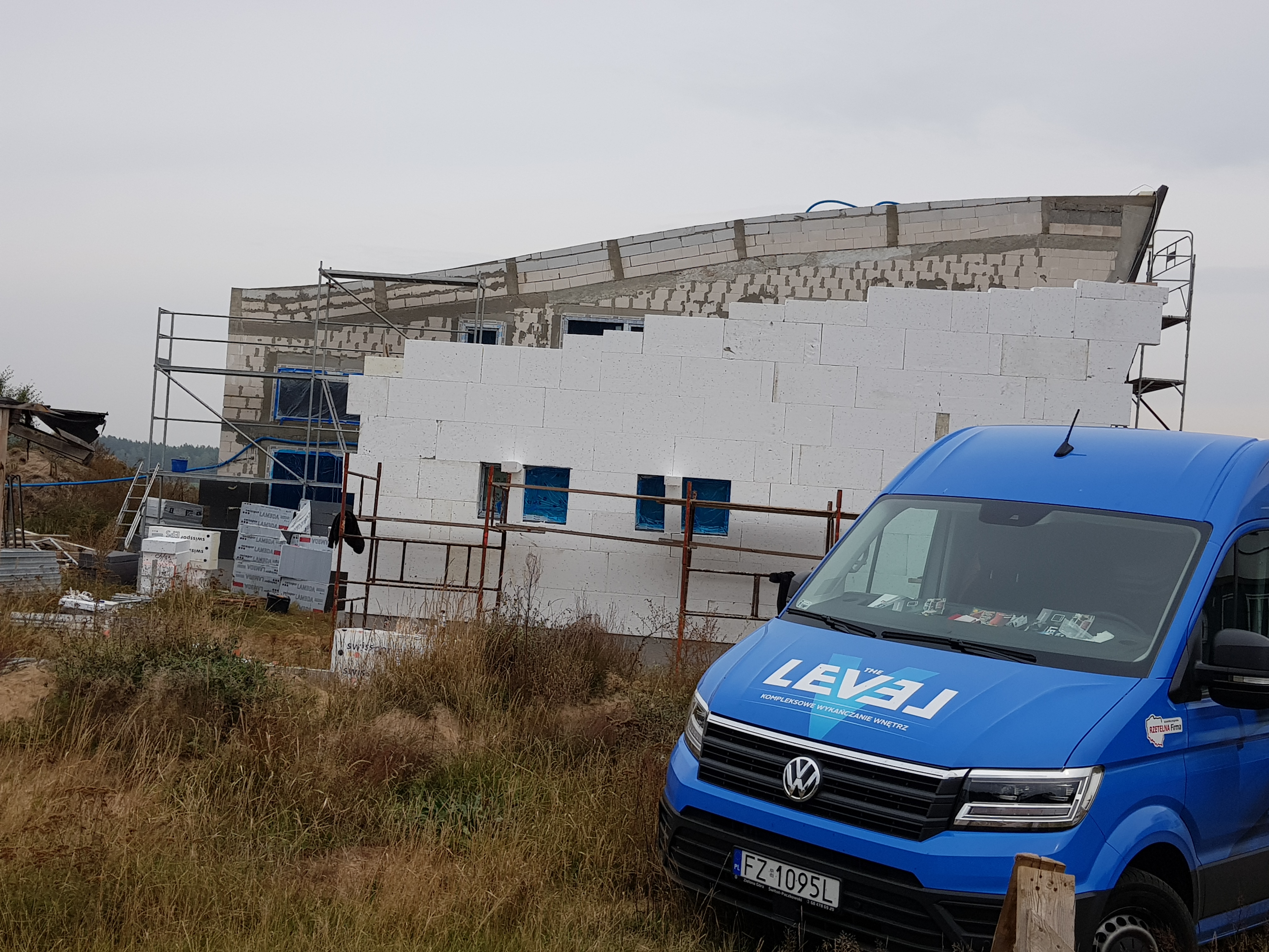 Budynek w trakcie budowy z białych bloczków, widoczne rusztowania i niebieski samochód dostawczy z logo firmy wykończeniowej na pierwszym planie.