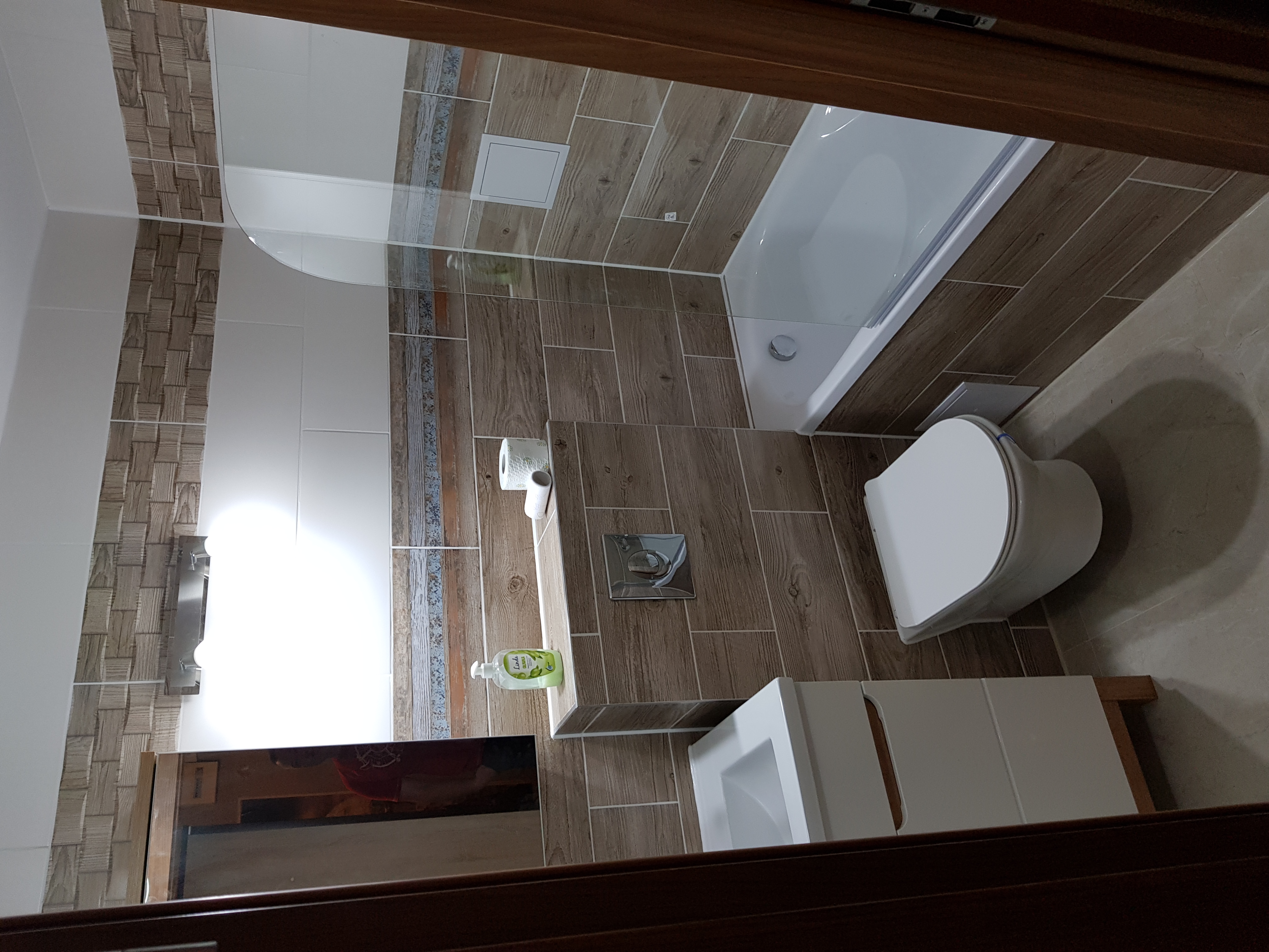 Nowoczesna łazienka z drewnopodobnymi płytkami, białą armaturą i minimalistycznym designem, widziana z perspektywy wejścia.