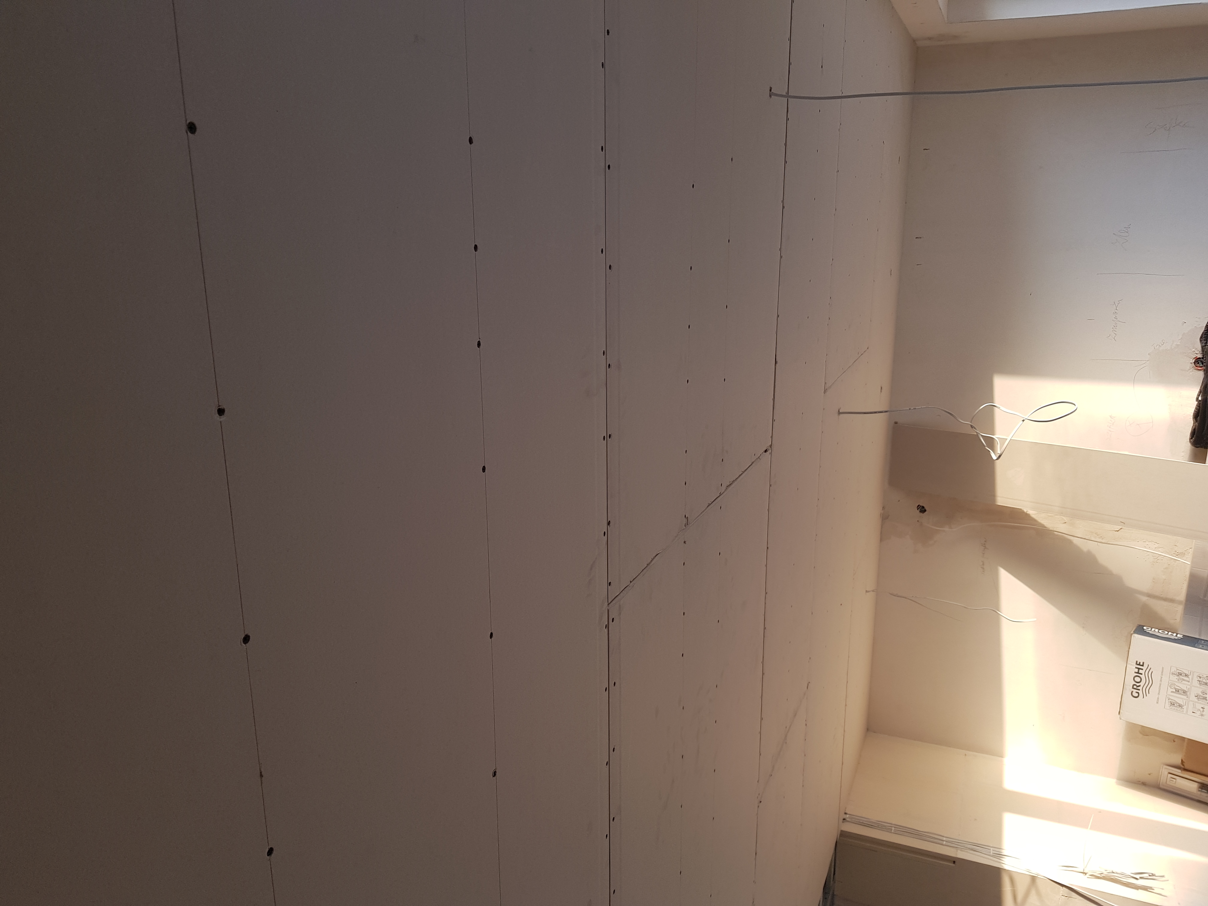 Ściana z płyt gipsowo-kartonowych, widoczne łączenia płyt i ślady po szpachlowaniu, przewody elektryczne wystające ze ściany, w tle karton Grohe.
