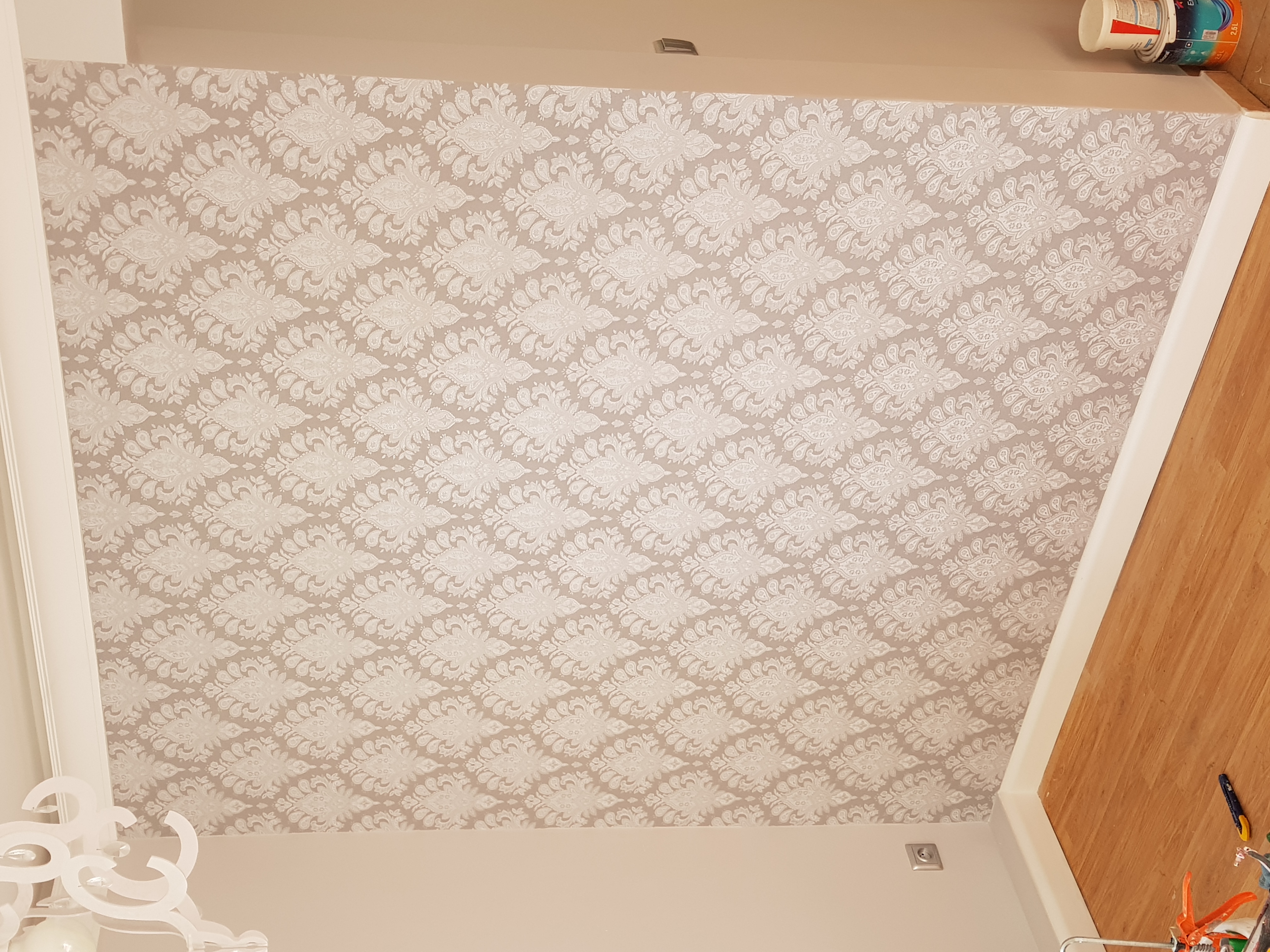 Beżowa tapeta w białe ornamenty na ścianie, widoczne puszki z farbą przy suficie i fragment podłogi z narzędziami, ujęcie z dołu