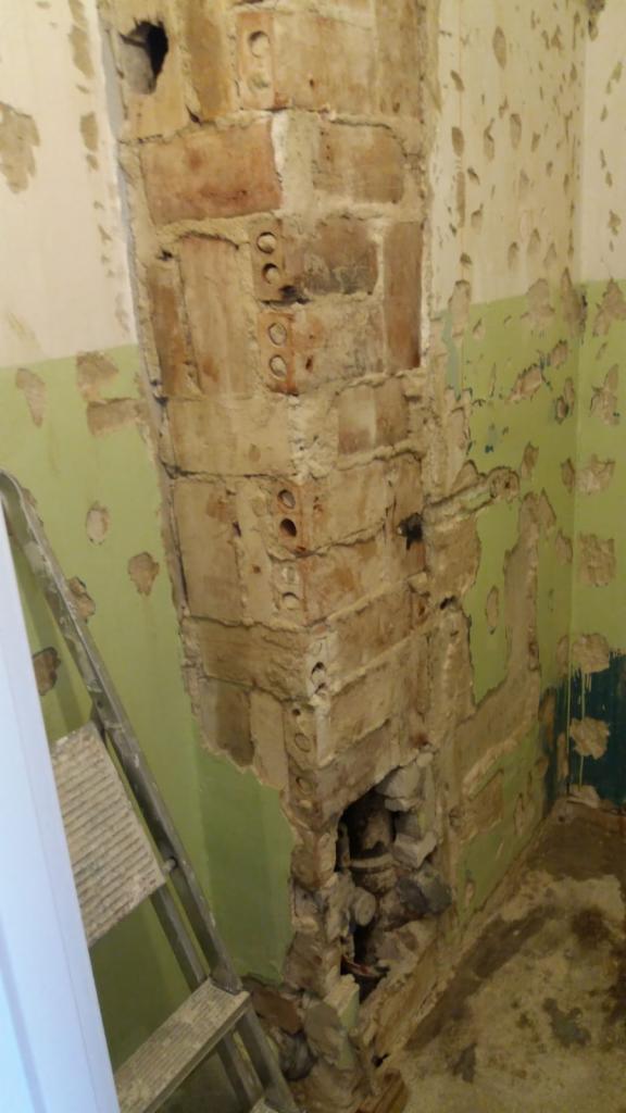 Demontaż fragmentu ściany z cegły w narożniku pomieszczenia z resztkami zielonej farby, widoczna drabina aluminiowa.