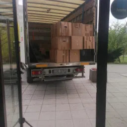 Otwarta paka samochodu dostawczego wypełniona kartonami z uchwytami, widoczny numer rejestracyjny, częściowo widoczne otoczenie budynku przez otwarte drzwi.