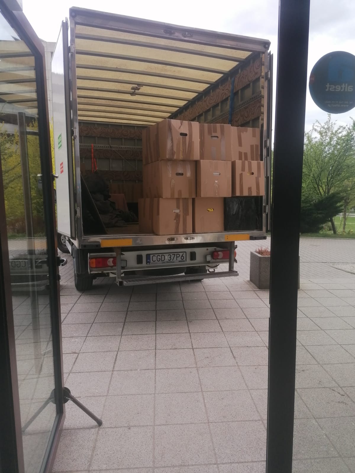 Otwarta paka samochodu dostawczego wypełniona kartonami z uchwytami, widoczny numer rejestracyjny, częściowo widoczne otoczenie budynku przez otwarte drzwi.