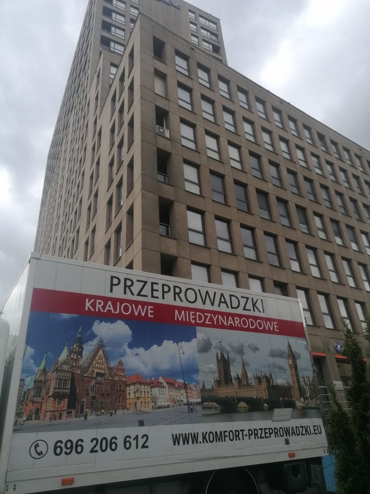 Reklama na samochodzie dostawczym z grafiką przedstawiającą Wrocław i Londyn, informująca o krajowych i międzynarodowych usługach transportowych na tle wysokiego biurowca.