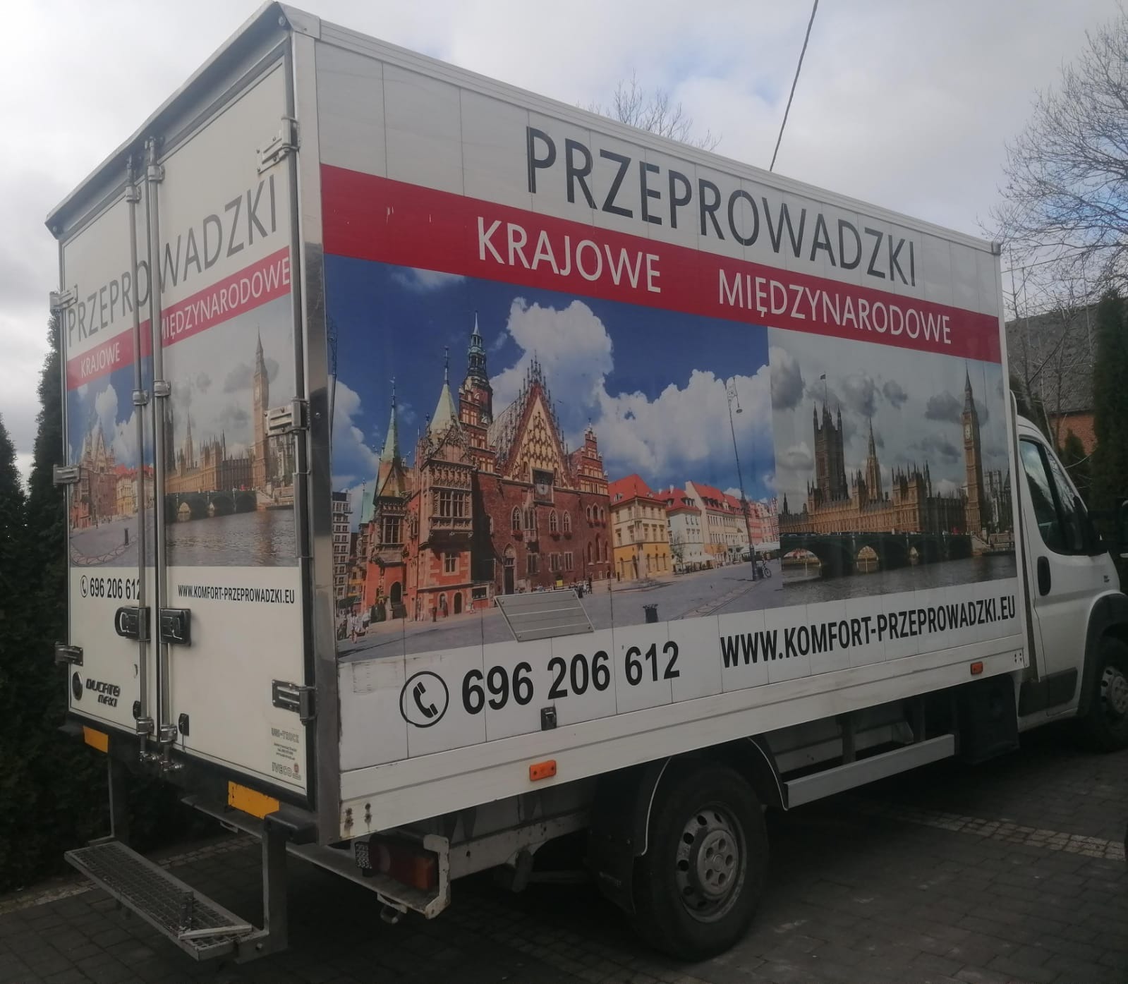 Biała furgonetka z grafiką przedstawiającą zabytki Wrocławia i Londynu oraz napisem 'Przeprowadzki krajowe i międzynarodowe' i numerem telefonu.