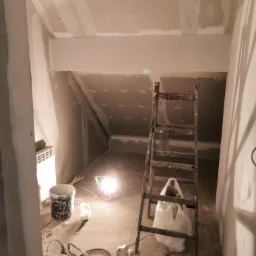Poddasze w trakcie remontu, widoczne płyty kartonowo-gipsowe na ścianach i suficie, drabina oparta o ścianę, wiadro z farbą oraz mieszadło do gładzi na podłodze, oświetlone lampą budowlaną.